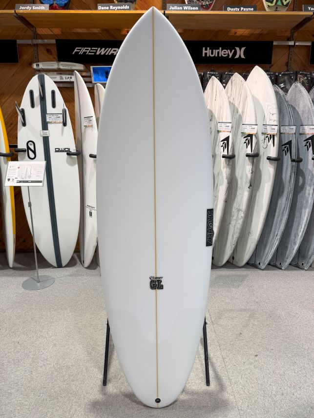 White surfboard on a display stand in a surf shop, with other boards and brand banners in the background.