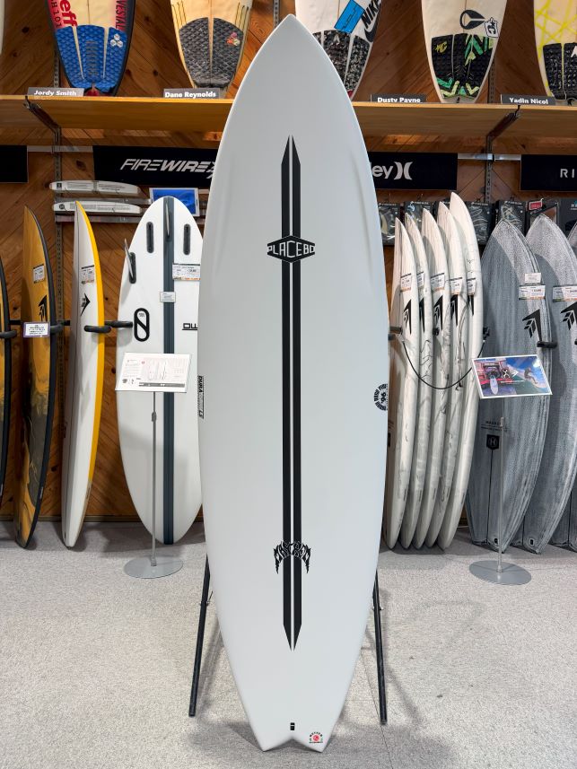 White surfboard with a black double stripe and Placebo logo on a stand in a surf shop, with other boards displayed behind.