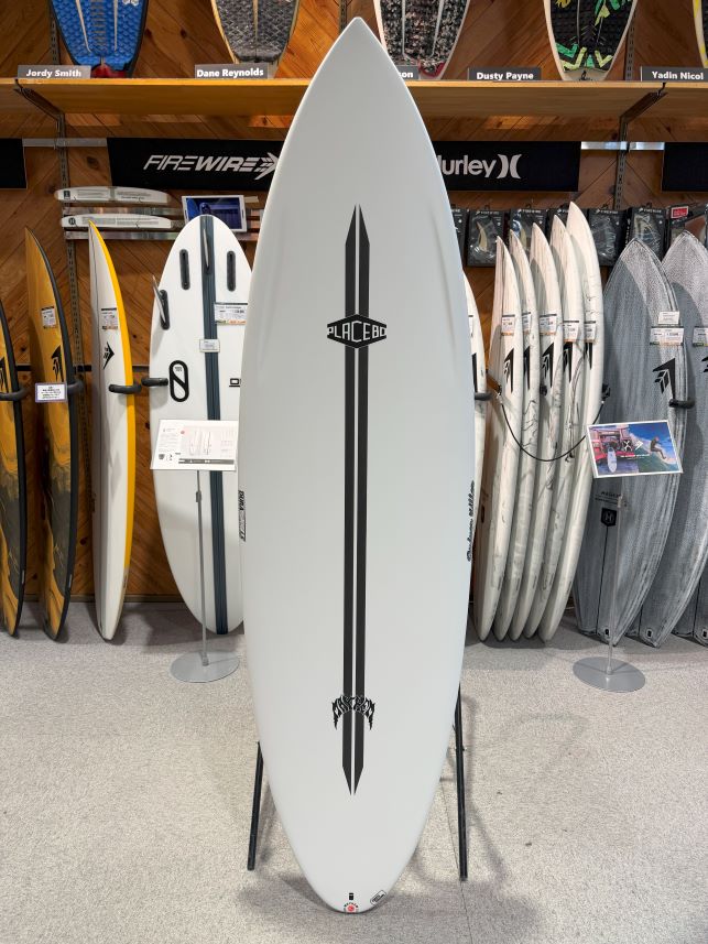 White surfboard on a display stand in a surf shop, featuring a bold black vertical stripe design and a Placebo logo down the center.