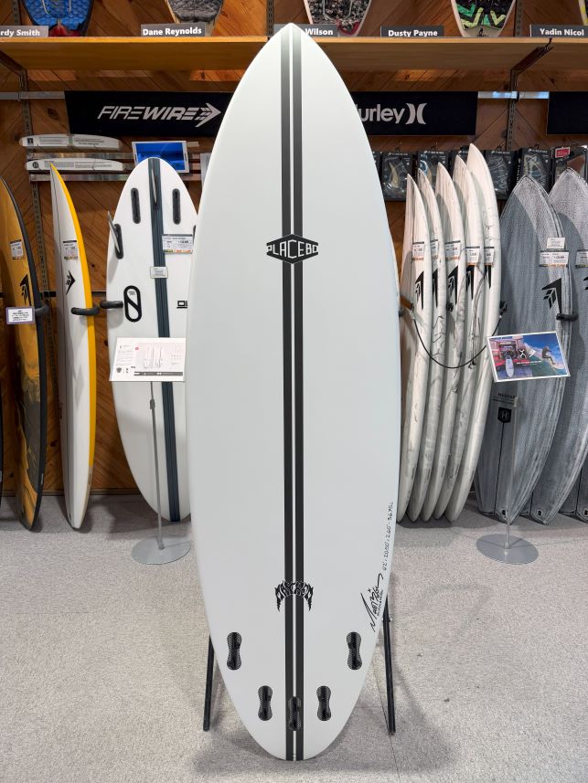 White surfboard with two black center rails and a Placebo logo on display in a surf shop, with other boards around.
