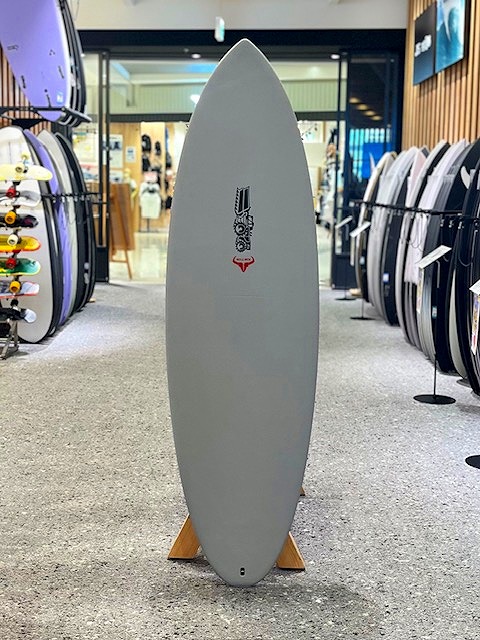a-IMG_3220 - SHOP NEWS | ムラサキスポーツオンラインストア 通販 Gray surfboard on a wooden stand in a surf shop, surrounded by other boards on racks inside a showroom.