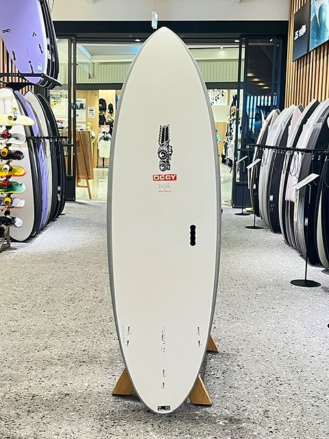 a-IMG_3225 - SHOP NEWS | ムラサキスポーツオンラインストア 通販 White surfboard standing upright on a wooden rack inside a surf shop, surrounded by other boards on racks.