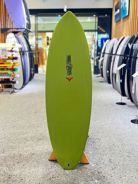 a-IMG_3243 - SHOP NEWS | ムラサキスポーツオンラインストア 通販 Green longboard surfboard standing upright on a wooden rack inside a surf shop, with other boards visible on shelves around it.