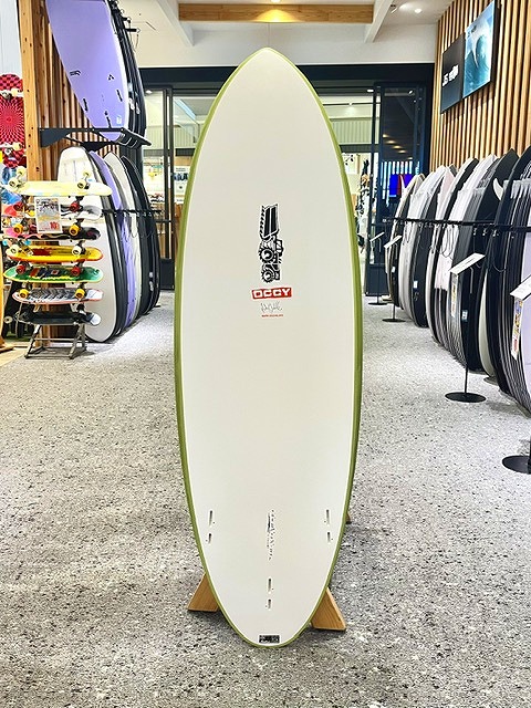 a-IMG_3248 - SHOP NEWS | ムラサキスポーツオンラインストア 通販 White surfboard with a lime-green edge on a wooden display stand in a surf shop, surrounded by other boards on racks.