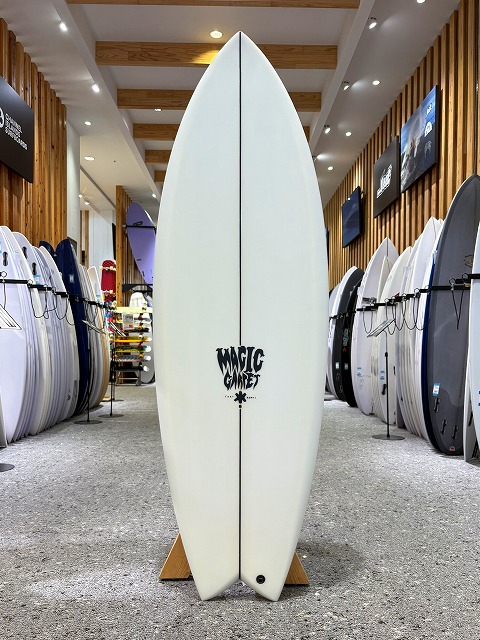 a-image0 - SHOP NEWS | ムラサキスポーツオンラインストア 通販 White surfboard standing upright on a wooden rack in a surf shop, with wood-paneled walls and many other boards on either side, featuring a black 'Magic' logo.