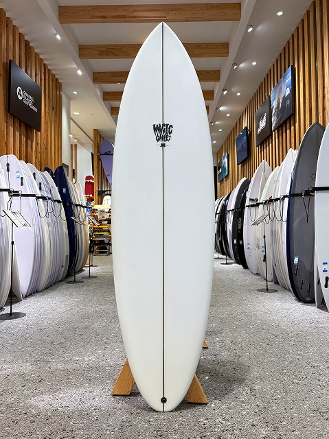 a-image0 - SHOP NEWS | ムラサキスポーツオンラインストア 通販 White surfboard standing upright on a wooden rack in a showroom, surrounded by other boards and wall-mounted displays in a surfing shop.