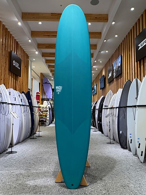 a-image0 - SHOP NEWS | ムラサキスポーツオンラインストア 通販 Tall turquoise surfboard standing upright on a wooden display stand inside a surf shop, surrounded by racks of other boards along the walls.
