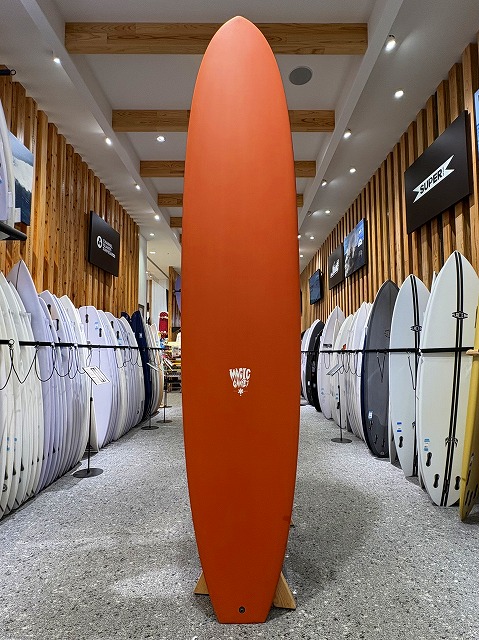 a-image0 - SHOP NEWS | ムラサキスポーツオンラインストア 通販 Orange surfboard standing upright on a showroom floor between rows of white and light-colored surfboards, with wooden wall panels and overhead lights in a surf shop.