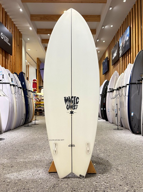 a-image1 - SHOP NEWS | ムラサキスポーツオンラインストア 通販 White surfboard with a 'Magic' logo standing upright on a wooden stand in a surf shop showroom. Surrounding racks hold numerous boards on both sides.
