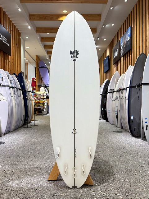 a-image1 - SHOP NEWS | ムラサキスポーツオンラインストア 通販 White surfboard standing upright on a display rack in a surf shop, with other boards lined up along the walls and wood-paneled ceiling in the background.