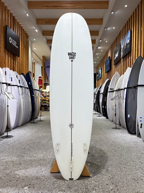 a-image1 - SHOP NEWS | ムラサキスポーツオンラインストア 通販 White surfboard upright on a wooden stand in a surf shop, with rows of other boards on racks along wood-paneled walls.