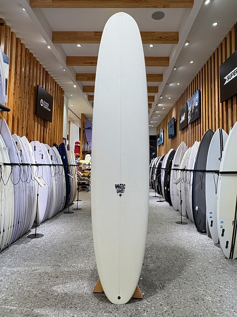 a-image1 - SHOP NEWS | ムラサキスポーツオンラインストア 通販 White longboard surfboard standing upright on a wooden display stand in a surf shop, flanked by many other surfboards along racks.