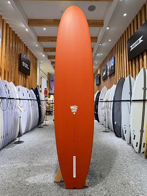 a-image1 - SHOP NEWS | ムラサキスポーツオンラインストア 通販 Tall orange surfboard displayed upright on a stand in a surf shop, flanked by white and colored boards along wooden walls.