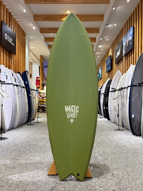 a-image2 - SHOP NEWS | ムラサキスポーツオンラインストア 通販 Green surfboard on a wooden stand in a surf shop showroom, centered with the 'Magic' logo, surrounded by rows of other boards on racks.