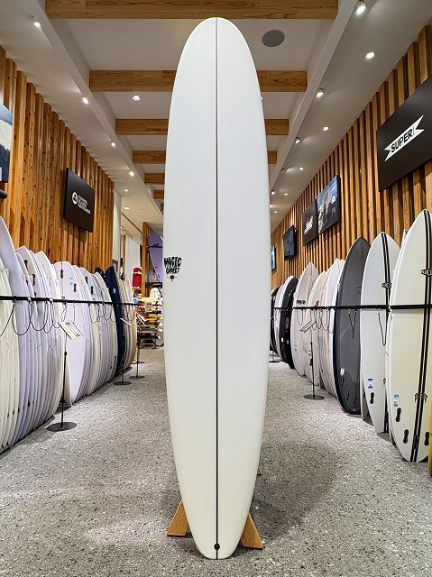 a-image2 - SHOP NEWS | ムラサキスポーツオンラインストア 通販 Tall white surfboard standing upright on a wooden stand in a surf shop showroom amid rows of boards on both sides.