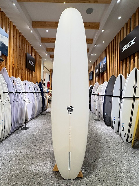 a-image2 - SHOP NEWS | ムラサキスポーツオンラインストア 通販 Tall white surfboard displayed upright in a surf shop, flanked by racks of boards along wooden walls and bright overhead lighting.