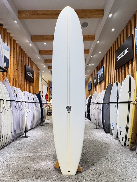a-image2 - SHOP NEWS | ムラサキスポーツオンラインストア 通販 White surfboard standing upright on a wooden rack in a surf shop aisle, with numerous boards lined along both walls.