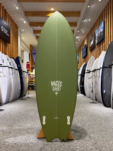 a-image3 - SHOP NEWS | ムラサキスポーツオンラインストア 通販 Olive green surfboard standing upright on a display rack in a shop, with rows of surfboards lined up along both walls.