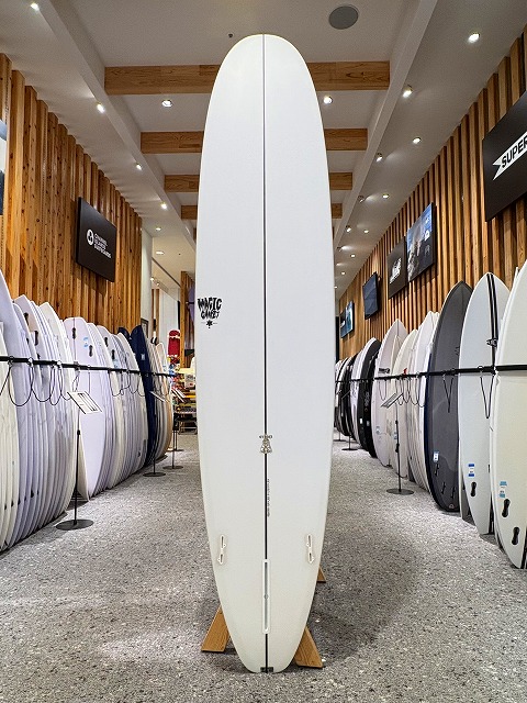 a-image3 - SHOP NEWS | ムラサキスポーツオンラインストア 通販 Tall white surfboard standing upright on a wooden stand in a surf shop, surrounded by rows of boards.