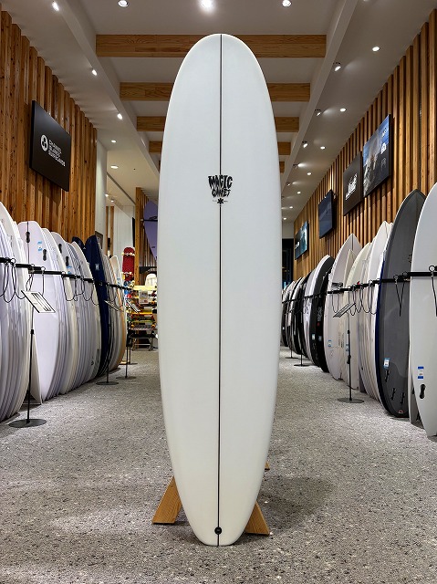 a-image6 - SHOP NEWS | ムラサキスポーツオンラインストア 通販 Long white surfboard standing upright on a wooden stand in a surf shop, with racks of boards lining both sides of the aisle.