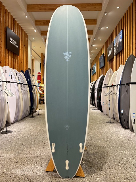 a-image9 - SHOP NEWS | ムラサキスポーツオンラインストア 通販 Tall blue-green surfboard on a wooden stand in a showroom, with numerous boards lining racks along the walls.