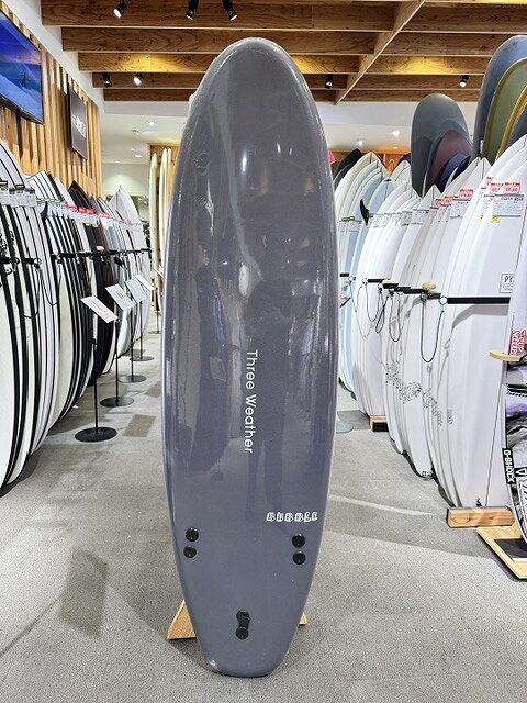 Tall gray surfboard standing upright on a stand in a surf shop, surrounded by racks of white and colored boards.