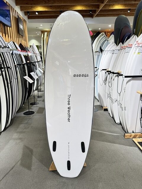 White surfboard with three fin boxes standing upright on a display rack in a surf shop, surrounded by numerous other boards; brand name 'Three Weather' visible along the center.