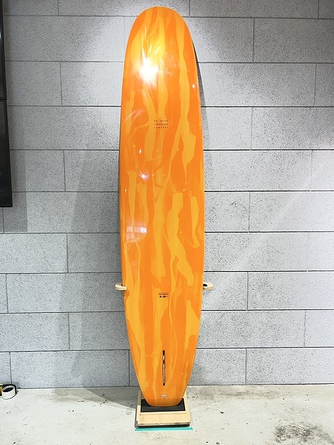 Orange longboard skateboard standing upright against a gray brick wall, resting on a small wooden base.
