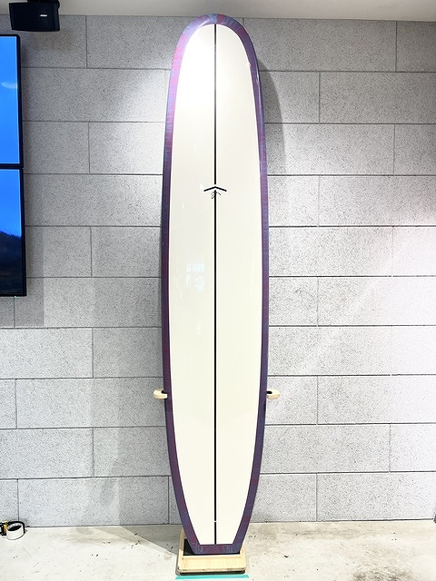 Tall surfboard standing upright on a wooden stand against a gray tiled wall; cream center with purple rails.