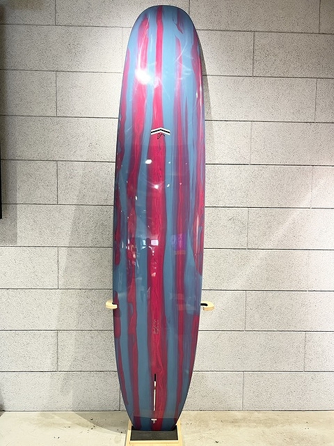 Colorful surfboard with pink and blue vertical stripes standing on a wall rack against a gray brick wall, logo visible in center.