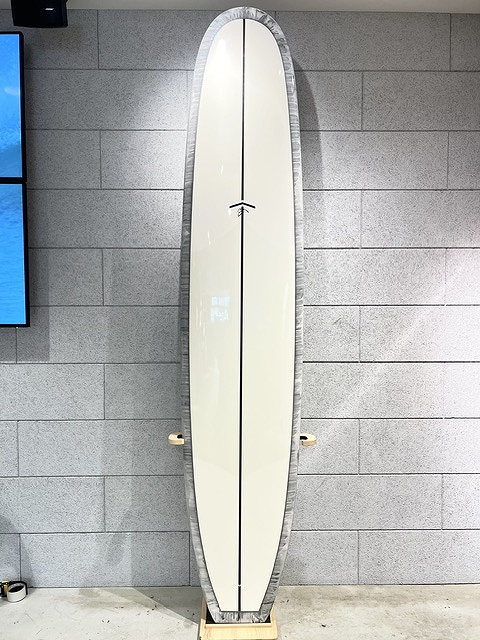 Cream-colored surfboard with a black center seam, standing upright on a wooden rack against a gray brick wall.