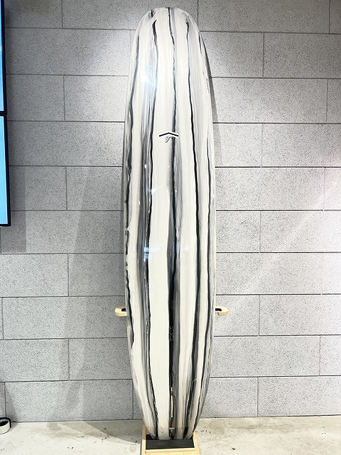 Tall white surfboard with black vertical stripes, standing upright on a wooden rack against a gray wall in a showroom setting.
