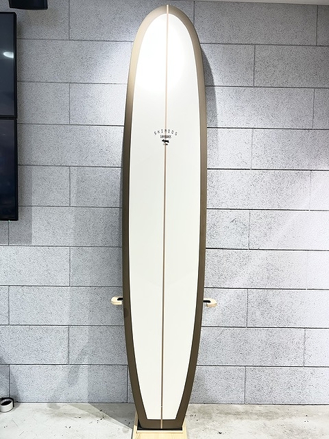 s-IMG_6379 - SHOP NEWS | ムラサキスポーツオンラインストア 通販 A white surfboard with a dark brown edge standing upright against a gray tiled wall on a wooden stand, no fins visible.