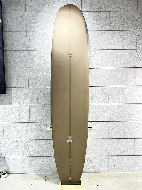 s-IMG_6380 - SHOP NEWS | ムラサキスポーツオンラインストア 通販 Brown longboard surfboard standing upright on a wooden display stand against a gray brick wall, center stringer visible down the board.