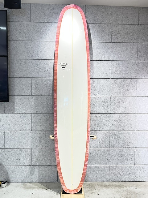 s-IMG_6381 - SHOP NEWS | ムラサキスポーツオンラインストア 通販 White surfboard with a pink border standing upright on a wooden rack against a gray tiled wall in an indoor display.