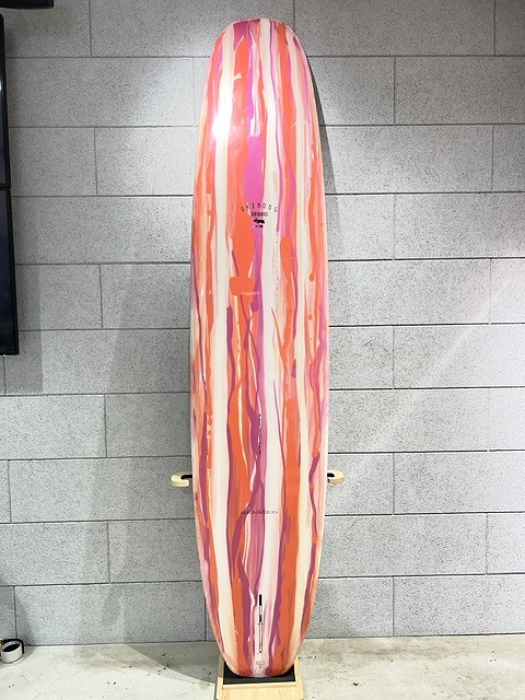 s-IMG_6382 - SHOP NEWS | ムラサキスポーツオンラインストア 通販 Pink, orange, and white striped surfboard standing upright against a gray brick wall on a wooden stand.