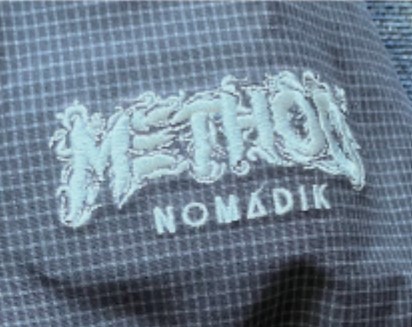 Close-up of blue checkered fabric with white embroidered logo; a stylized word above the word 'NOMADIK'.
