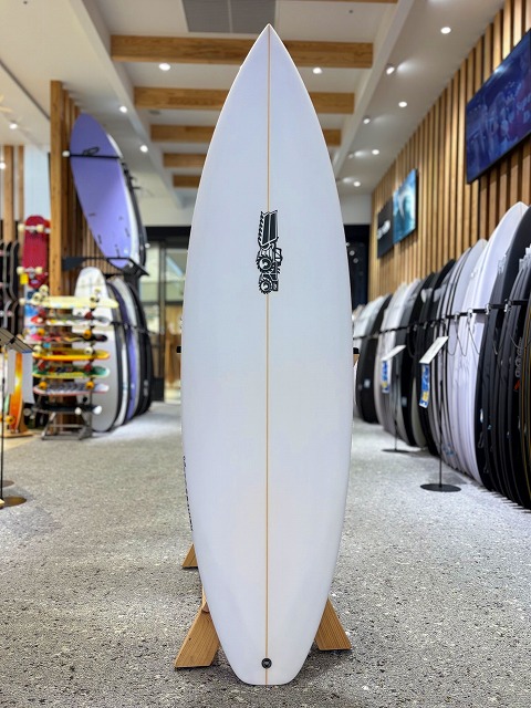 ゼログラビティー 1 - SHOP NEWS | ムラサキスポーツオンラインストア 通販 White surfboard on a wooden stand in a surf shop, logo mid-board with racks of boards visible in the background on both sides.