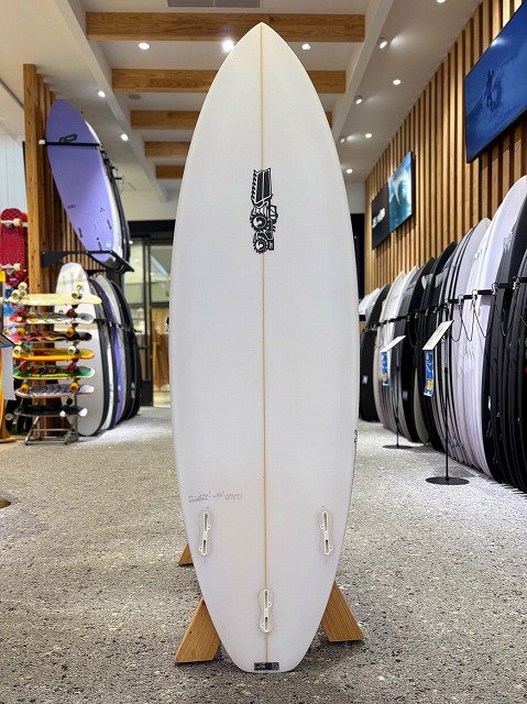 ゼログラビティー 2 - SHOP NEWS | ムラサキスポーツオンラインストア 通販 White surfboard standing upright on a wooden display rack inside a surf shop, with other boards lining the walls.