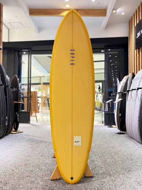 マージン 0 - SHOP NEWS | ムラサキスポーツオンラインストア 通販 Yellow surfboard on a wooden stand, displayed upright in a shop surrounded by other boards.