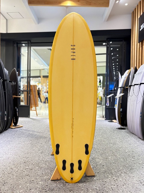 マージン 1 - SHOP NEWS | ムラサキスポーツオンラインストア 通販 Yellow surfboard standing upright on a wooden stand in a surf shop, with other boards on display in the background.