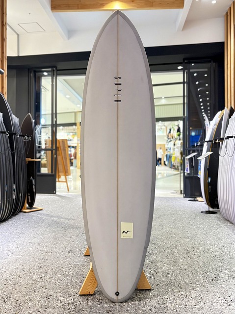 マージン 2 - SHOP NEWS | ムラサキスポーツオンラインストア 通販 White surfboard on a wooden stand in a surf shop, with other boards on display in the background.