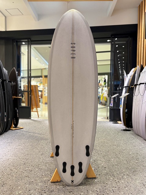 マージン 3 - SHOP NEWS | ムラサキスポーツオンラインストア 通販 Light-colored surfboard standing upright on display racks in a surf shop, with multiple fin boxes visible along the bottom and other boards in the background.