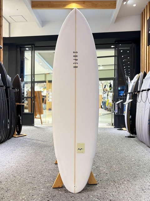 マージン 4 - SHOP NEWS | ムラサキスポーツオンラインストア 通販 White surfboard on a wooden stand centered in a surf shop, with other boards lined up along the sides.