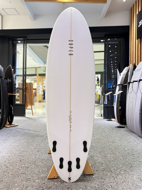 マージン 5 - SHOP NEWS | ムラサキスポーツオンラインストア 通販 White surfboard on a display stand inside a surf shop, with other boards visible in the background.