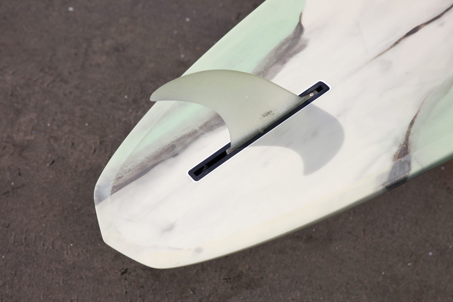 Hires_HI__IMG_4338-1536x1024 - SHOP NEWS | ムラサキスポーツオンラインストア 通販 Close-up of a light-colored surfboard tail with a translucent fin in the slot on a dark surface.