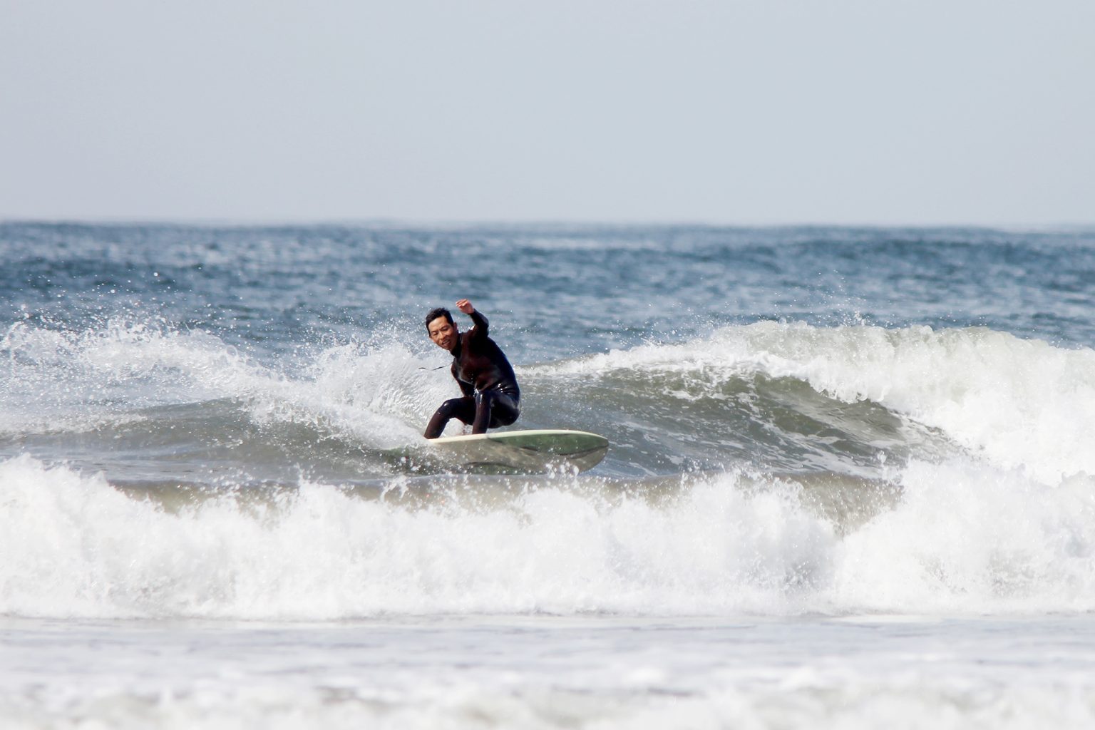 Hires_HI__RU_IMG_4710-1536x1024 - SHOP NEWS | ムラサキスポーツオンラインストア 通販 Surfer wearing a black wetsuit rides a wave on a white surfboard near the shore, spray flying around him.