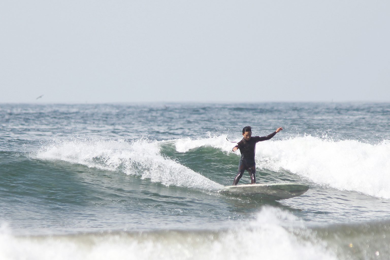 Hires_HI__RU_IMG_4834s-1536x1024 - SHOP NEWS | ムラサキスポーツオンラインストア 通販 Surfer standing on a board riding a small wave near the shore, wearing a black wetsuit and extending one arm for balance.