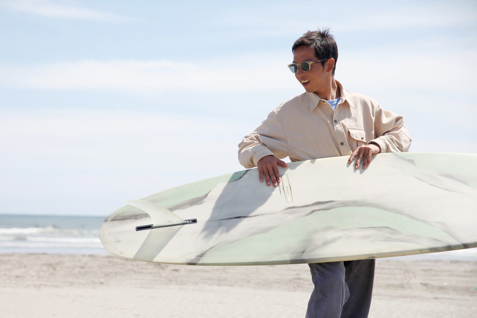 Hires_HI__snap_IMG_5959-1536x1024 - SHOP NEWS | ムラサキスポーツオンラインストア 通販 Man at the beach wearing sunglasses and a beige shirt, carrying a surfboard under his arm with the ocean in the background.