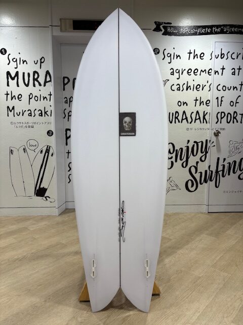 Two white surfboards standing upright on a wooden rack inside a surf shop, with black surfing-themed wall art; center board has a small skull sticker.
